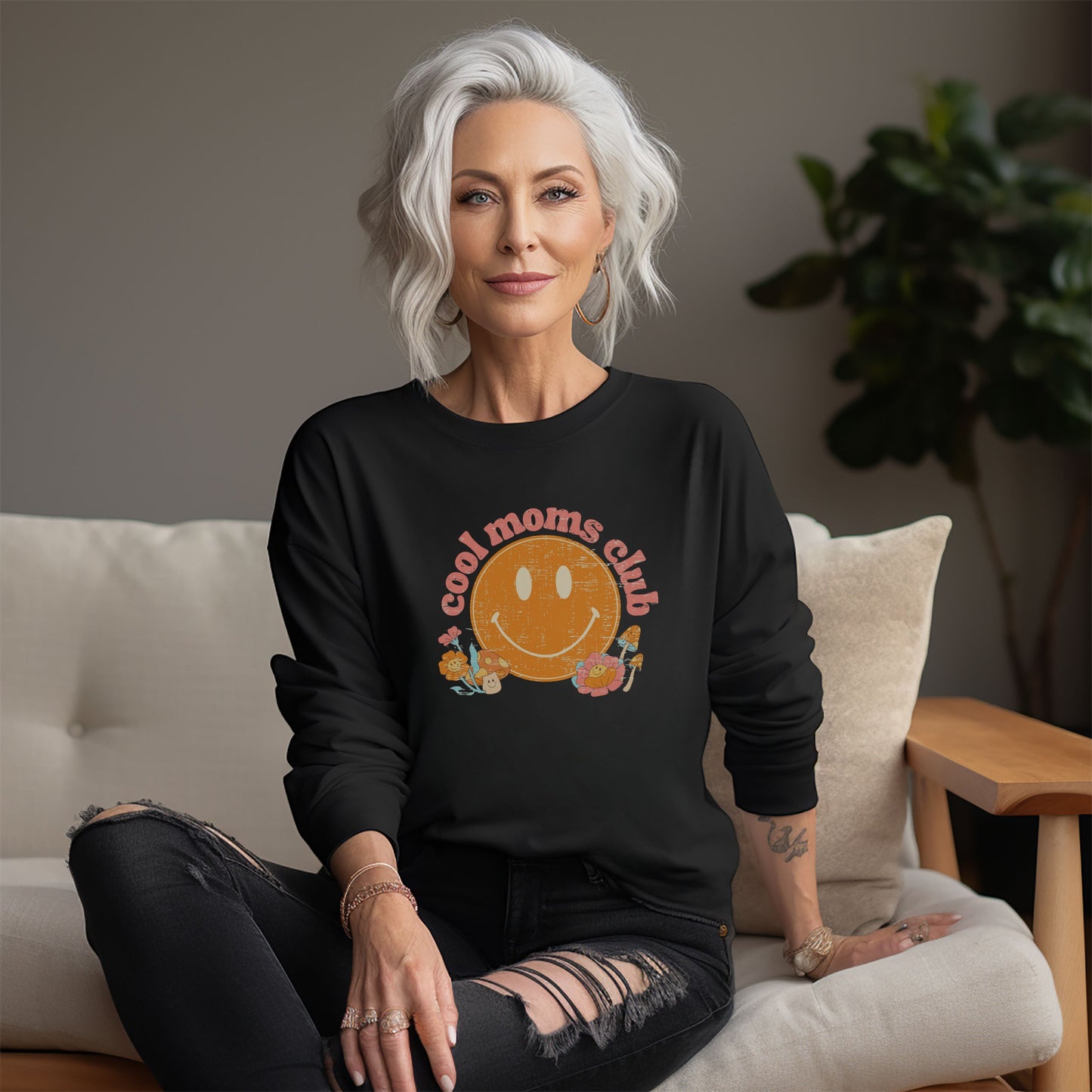 Woman with white hair wearing a black crewneck sweatshirt with a retro smiley face and mushrooms with vintage pink font that reads: “cool moms club”. Fun Mother’s Day Gift!