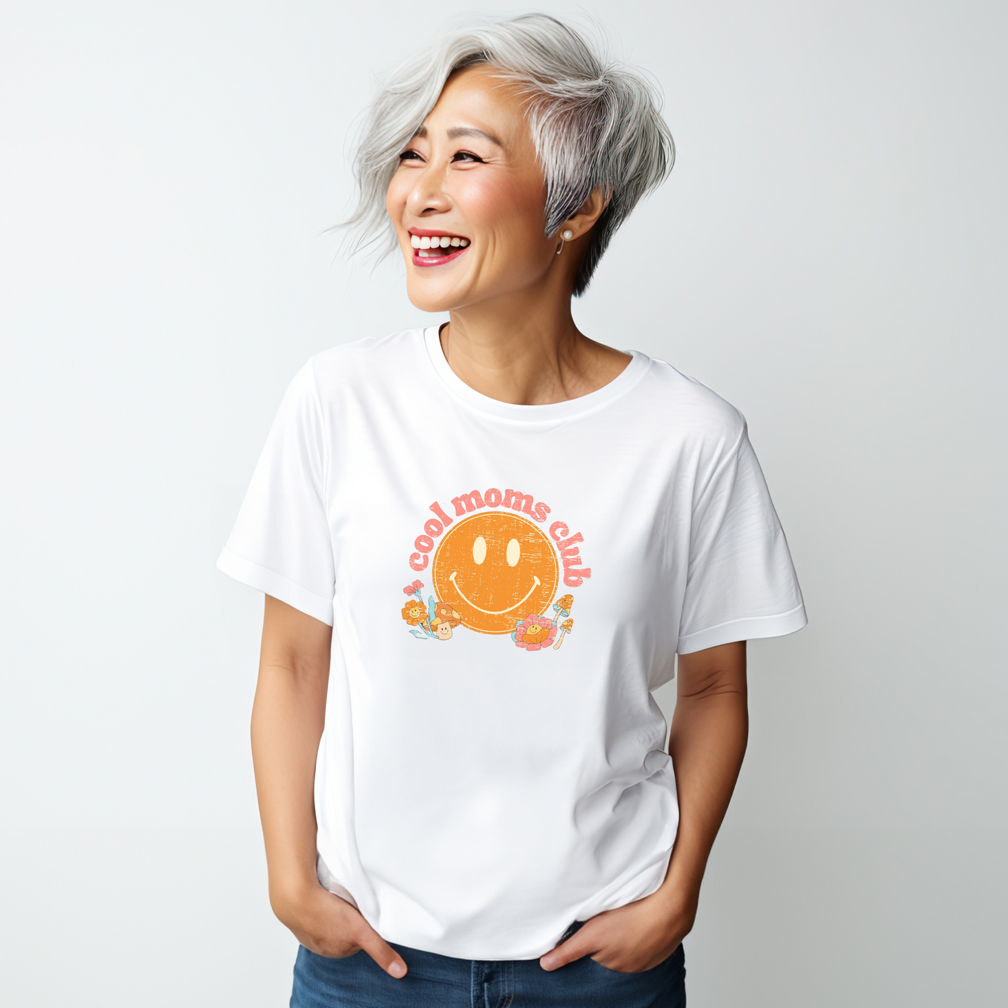 Asian woman with grey hair smiling while wearing a white t-shirt that says “cool moms club” in a vintage pink font with a retro smiley face and mushrooms.