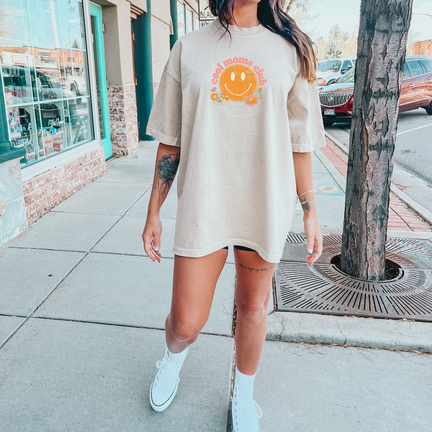 Woman wearing a natural color t-shirt that says “cool moms club” in a vintage pink font with a retro smiley face and mushrooms.