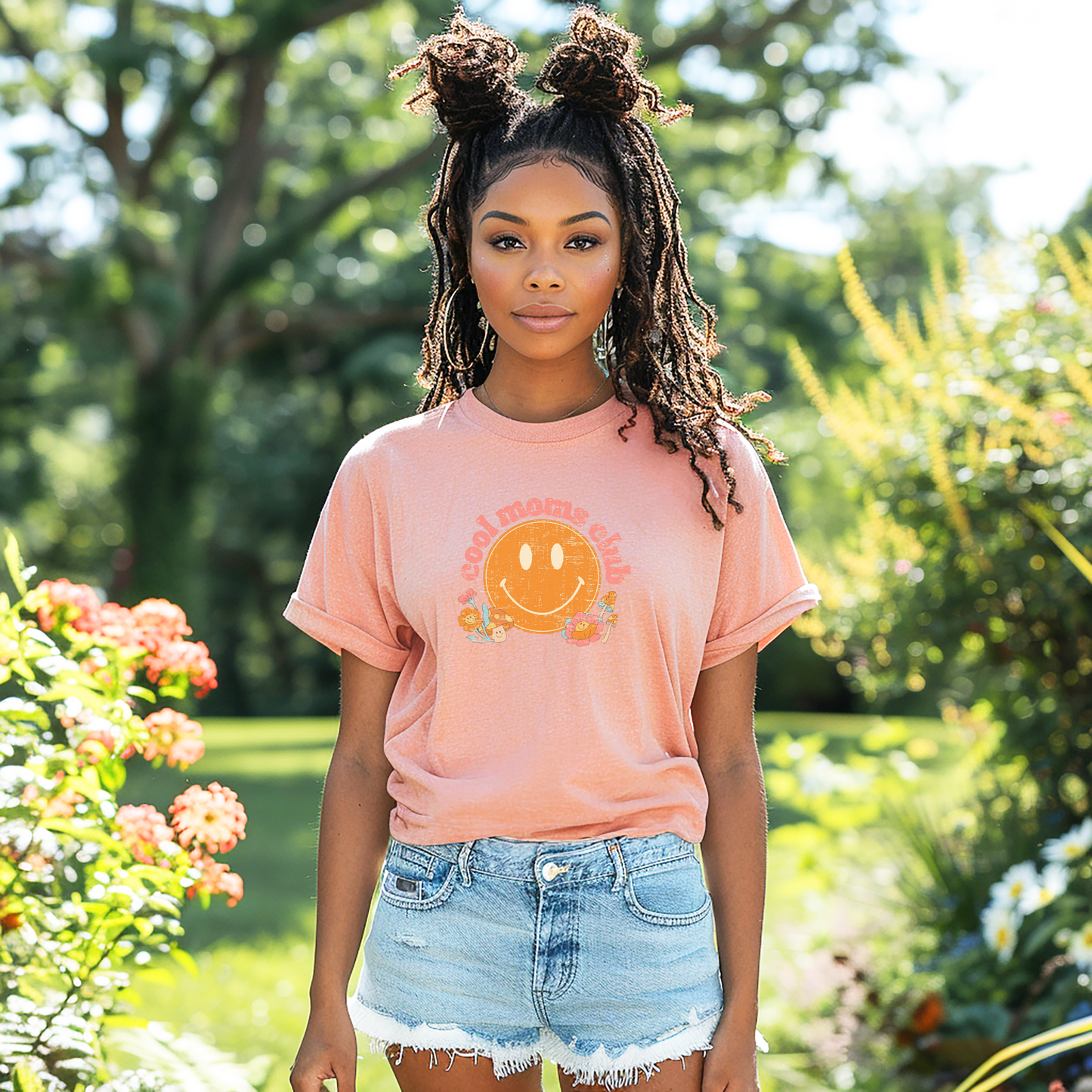 Black woman standing in a garden wearing a heather peach t-shirt that says “cool moms club” in a vintage pink font with a retro smiley face and mushrooms.