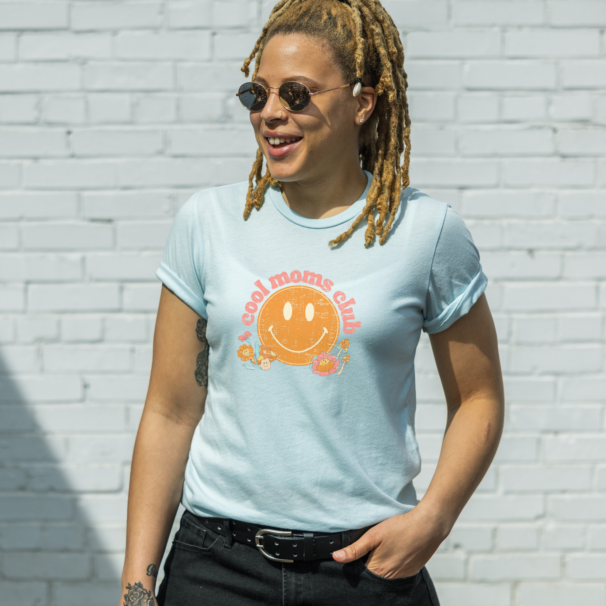 Black woman with dreadlocks smiling while wearing a heather ice blue t-shirt that says “cool moms club” in a vintage pink font with a retro smiley face and mushrooms.