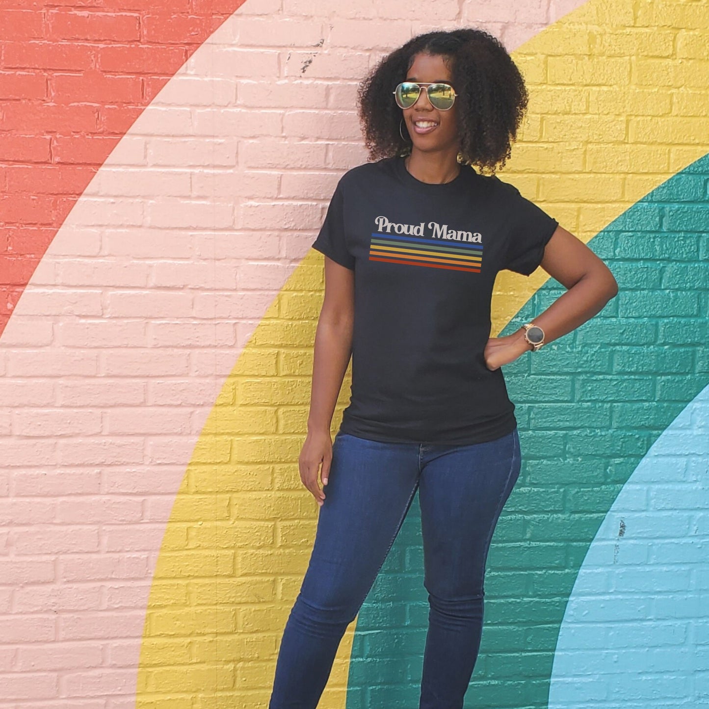 Black woman standing in front of rainbow wall wearing a black “Proud Mama” rainbow t-shirt. 